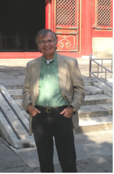Ron Schleifer has short gray hair and glasses and is smiling. He is wearing a green collared shirt, a beige blazer, and dark blue jeans. He is standing outside. In the background is a building with concrete steps leading to a red door entryway.